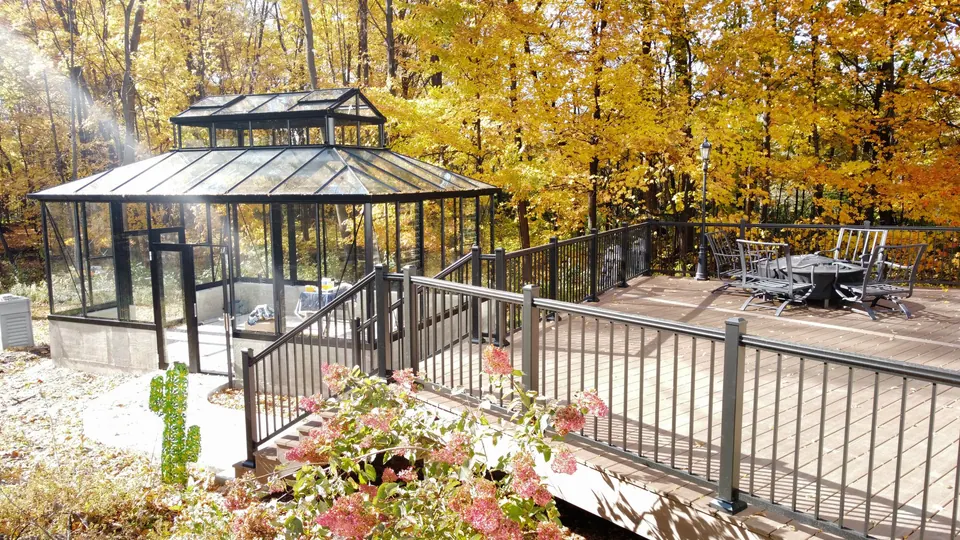 Finished composite deck with black aluminum railing and a glass-walled gazebo on a white two-story house among fall trees